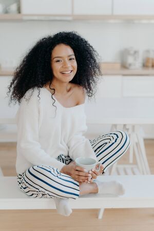 Image of lovely curly haired lady drinks coffee or tea from white mug, wears fashionable white sweater, striped pants, poses at kitchen against blurred background. People and lifestyle conceptの写真素材