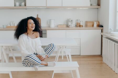 Horizontal shot of overjoyed dark skinned woman laughs pleasantly, drinks coffee, looks out of window in kitchen, dressed in fashionable clothes. Smiling lady with hot tasty beverage, relaxes at homeの写真素材