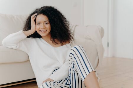 Lovely dark skinned woman with crisp hair, sits on floor near sofa, wears white sweater and striped trousers, enjoys positive emotions, enjoys weekend at home. People, rest, happiness conceptの写真素材