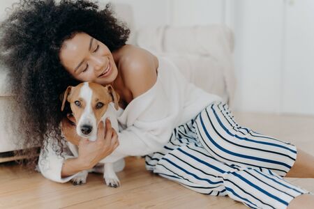Affectionate young woman hugs dog with love and care, keeps eyes closed from pleasure, smiles gently, has healthy dark skin, poses on floor, petting animal. People, friendship and pets conceptの写真素材