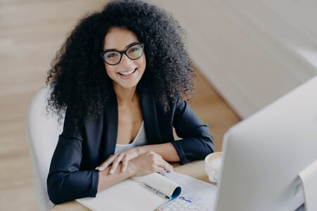 Top view of glad African American businesswoman smiles pleasantly, notes information, makes notes, wears transparent glasses, formal clothes, computer monitor in front, sits at desktop aloneの写真素材