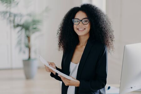 Photo of happy female entrepreneur with Afro hairstyle, studies documentation, wears spectacles and elegant clothes, stands in office interior, prepares to present her business ideas for colleaguesの写真素材