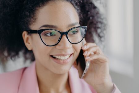 Headshot of beautiful smiling dark skinned woman wears optical glasses, holds modern cell phone, has...