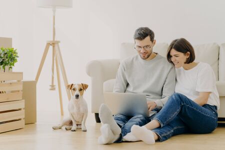 Beautiful couple with dog sit on floor in spacious living room, make purchasing online, buy furniture for new home, have to unpack belongings, have happy expression, start living together in new flatの写真素材