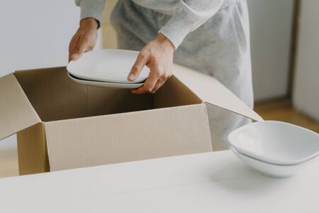 Cropped image of unknown faceless busy man unpacks boxes with dishes, holds white plates, moves in new apartment, tidies up at kitchen, has moving day. Bring out new plates. Relocation conceptの写真素材