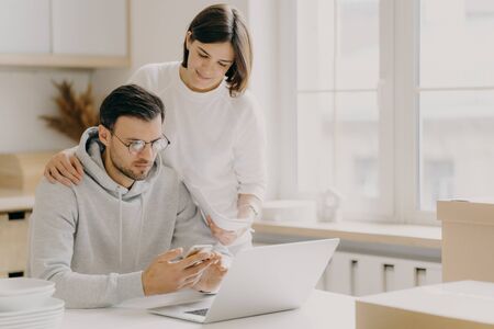 Picture of husband and wife study notification from bank, holds mobile phone and papers, work on laptop computer, pose in kitchen during relocation day, dressed in casual wear, save family moneyの写真素材