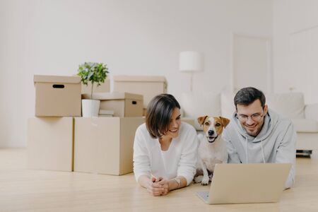 Family couple take break in their new apartment after moving in, lies on floor and surf internet via...