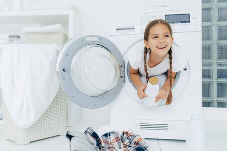 Happy European girl with two plaits, poses inside of wash machine, holds white bottle with liquid powder, has thoughtful expression, pile of dirty clothes in basin, looks somewhere into distanceの写真素材