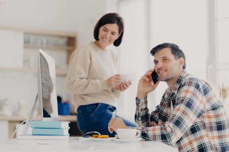 Photo of cheerful busy male freelancer uses mobile phone, discusses latest news, sits at desktop with papers and modern computer. Positive woman dressed in sweater and jeans, holds mug of drinkの写真素材