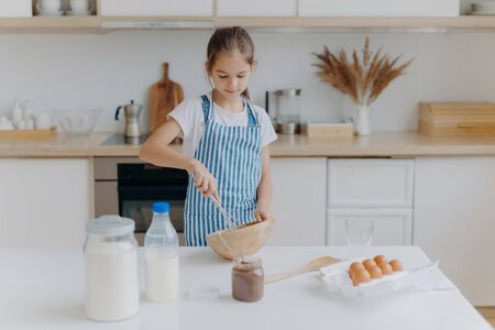 Small cute girl in apron, mixes ingredients, whisks with beater, uses eggs, milk, flour, tries new recipe, stands against kitchen interior, prepares tasty cookies or bakery, learns how to cook.の写真素材