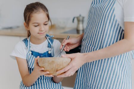 Busy little girl hold bowl, looks how mother whisks ingredients, learns how to cook. Unrecognizable mother in striped apron spends time with daughter, shows how to prepares dough. Cooking timeの写真素材