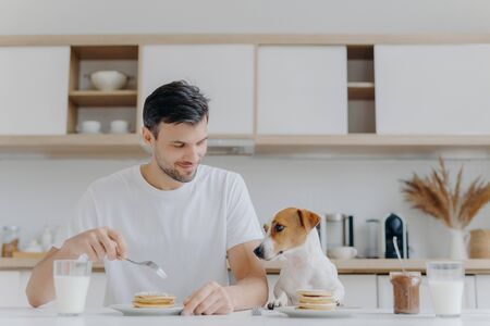 Photo of happy man eats tasty pancakes with fork, drinks milk from glass, wears white t shirt, his jack russell terrier dog poses near, has delicious breakfast together with host, kitchen backgroundの写真素材