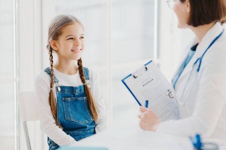 Happy little child listens attentively doctors advice and prescription, comes to see pediatrician in clinic, has two pigtails, wears denim overalls, talk about health problems, has checkup examinationの写真素材
