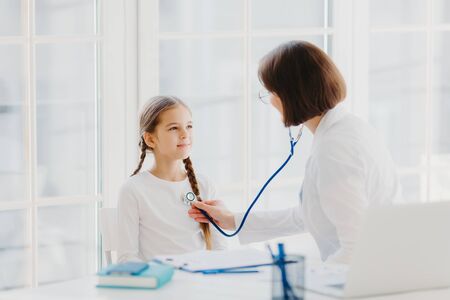 Woman pediatrician gives consultation to small girl, talk about health and symptoms, listens heart with phonendoscope, makes prescription, pose in hospital office. Childrean medical insurance and careの写真素材
