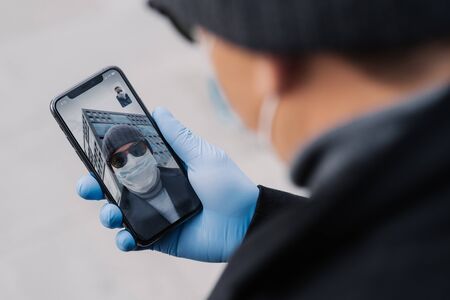 Male holds phone in medical gloves, talks with friend via online conference, greets friend, discuss latest news about coronavirus in their country. Medicine, health care and video consultationの写真素材