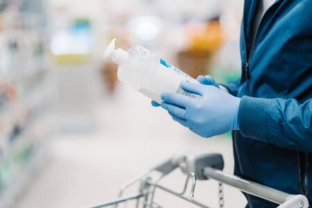 Horizontal shot of faceless man wears medical protective gloves during coronavirus outbreak, holds bottle of soap, cares about hygiene and disinfection during pandemic, makes shopping in storeの写真素材