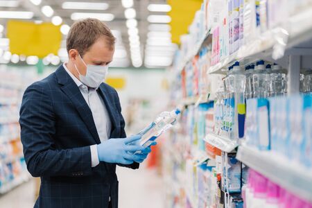 Photo of man dressed in formal suit, covers mouth with medical mask, wears rubber gloves to prevent virus spreading, poses in supermarket in washing agent department, holds household detergentの写真素材