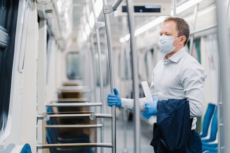 Virus hygiene and safety concept. Serious man dressed in elegant outfit, disposable mask and rubber gloves, touches handrail in underground carriage, commutes to office during quarantine timeの写真素材