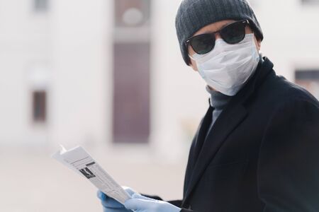 Coronavirus, health care, protection and quarantine concept. Serious man dresses hat, sunglasses, disposable medical mask and rubber gloves, concentrated somewhere, reads newspaper, poses outdoorの写真素材