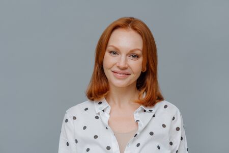 Portrait of attractive ginger European woman smiles gently at camera, wears makeup, dressed in polka...