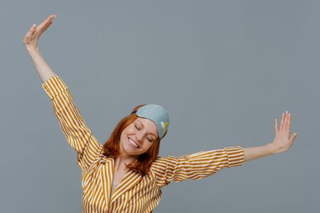 Perfect rest and sleeping concept. Cheerful young ginger European woman spreads arms and stretches after awakening, had sweet dreams, wears striped pajama and sleepmask, isolated on grey wallの写真素材