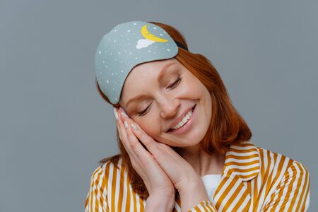 Close up shot of lovely tender woman has ginger hair, minimal makeup, leans at palms pressed together, smiles positively, wears blindfold on head, sees pleasant dreams. Healthy lifestyle conceptの写真素材