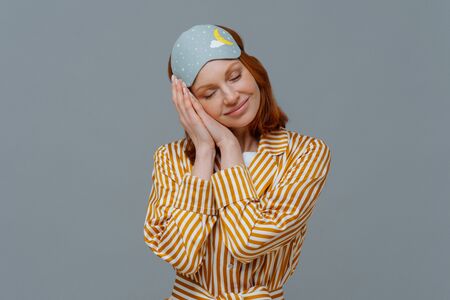 Pleased cheerful woman wears blindfold and striped pajamas, has happy expression, awakes in good...
