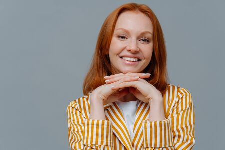 Close up shot of lovely woman has foxy hair, smiles toothily at camera, keeps hands under chin, dressed in homewear, enjoys calm domestic atmosphere, isolated on grey wall with blank space leftの写真素材