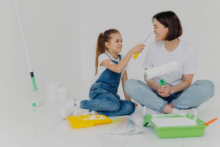 Positive girl smearks mums nose with paint by roller, have fun while making house renovation, sit together on floor in white room, use necessary tools for repairment. House renovation conceptの写真素材