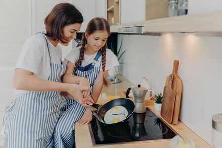 Busy mother and daughter wear striped aprons, pose at kitchen near cooker, fry eggs on pan, prepare fast breakfast, enjoy domestic atmosphere. Mum teaches small kid to cook. Happy family conceptの写真素材