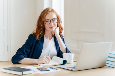 Redhead curly woman office worker analyzes data, makes accounting report, poses in coworking office, improves online sales, drinks coffee, wears spectacles and formal wear. Female employee indoorの写真素材