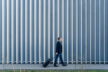 Horizontal panoramic shot of man passenger arrives in own country because of quarantine and pandemic situation in world, walks with suitcase, poses outdoor against metal fence, wears face maskの写真素材