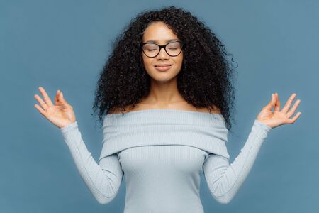Relaxed beautiful woman with Afro hairstyle, feels relieved, shows zen gesture, keeps hands in okay gesture, controls emotions, gets in control with herself has eyes closed isolated on blue backgroundの写真素材