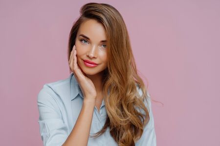 Studio shot of pleasant looking relaxed female model with long hair touches cheek gently, wears elegant shirt, looks straightly at camera, shows her beauty. Pretty businesswoman prepares to workの写真素材