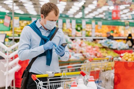 Young man poses in grocery store during coronavirus pandemic, wears protective medical mask and gloves, stands in supermarket near trolley, checks something in smartphone. Health, safety, quarantineの写真素材