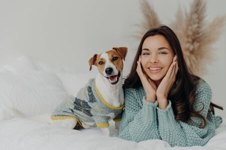 Horizontal shot of lovely brunette female in casual wear, lies on white bedclothes with pedigree dog, expresses happiness, enjoys company of pet, rest together at home, feel relaxed and satisfiedの写真素材
