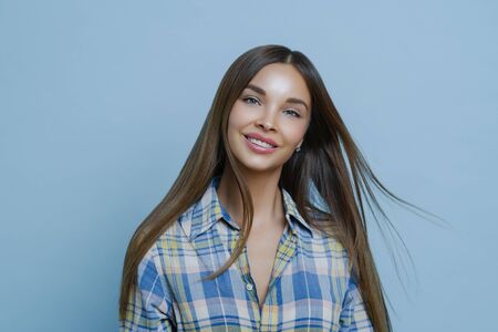 Charming pleased young woman smiles toothily, dressed in checkered shirt, has long straight dark...