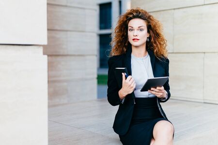 Horizontal portrait of pretty confident businesswoman dressed formally, holding her pocket book with pen and tablet computer, being busy with her work. People, lifestyle, career, business conceptの写真素材