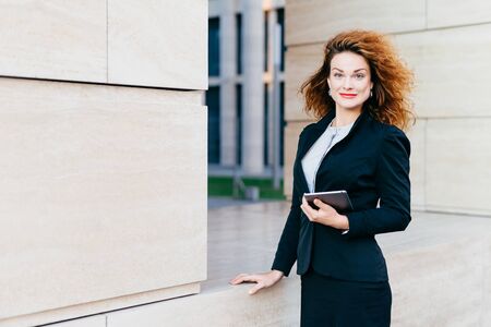 Pleasant-looking female dressed formally, holding digital tablet computer which is necessary for her work. Beatiful woman with curly bushy hair, having attractive appearance, going for her workの写真素材
