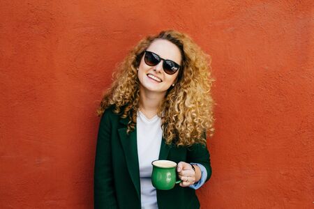 Medium close-up of pleased pretty woman with curly hair wearing sunglasses and jacket holding cup of cappuccino looking with smile into camera posing against orange studio wall. People and emotionsの写真素材