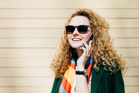 Young smiling office worker in sunglasses having blonde curly hair talking on cell phone during a break standing at street against beige background with copy space for your promotional contentの写真素材