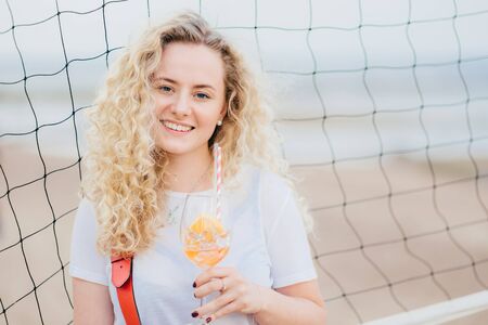 Adorable glad young European woman has bushy curly hair, smiles gently, holds glass of orange cocktail, stands near tennis net with copy space right for your advertisement or promotional textの写真素材