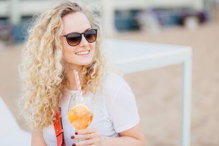 Positive blonde female with curly hair, wears sunglasses, looks with happy expression aside, has gentle smile, holds fresh cocktail, waits for friend on beach. People, resort, recreation conceptの写真素材