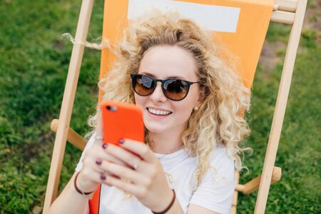 Positive European female with friendly smile, uses modern electronic device, happy to recieve message from boyfriend, rests outdoor during summer vacation. People, recreation, lifestyle conceptの写真素材