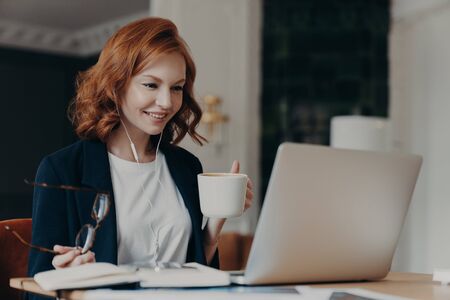 Young female teacher conducts lesson online, talks wih pupils via laptop computer, organizes video conference, teaches students, drinks aromatic coffee, holds glasses in hand, dressed formallyの写真素材