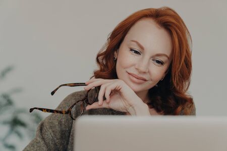 Photo of redhead woman takes off glasses, enjoys free time for e learning, focused at monitor of laptop computer, does distance job. Female student prepares for exams, uses modern technologiesの写真素材