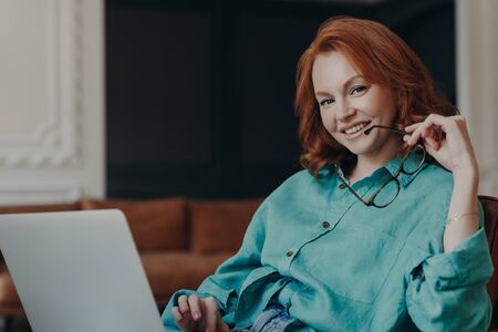 Positive good looking young woman freelancer works remotely on laptop computer, connected to wireless free internet at home, holds glasses, dressed in shirt, smiles happily, being in good moodの写真素材