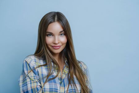 Portrait of good looking female student poses against blue studio wall, looks at camera with...