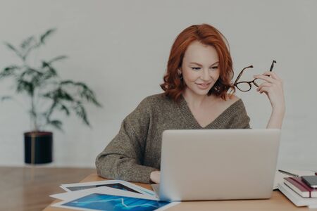 Busy redhead adult woman does freelance work on laptop computer, focused in monitor, prepares project, surrounded with paper documents, wears warm sweater, analyzes information, uses internetの写真素材