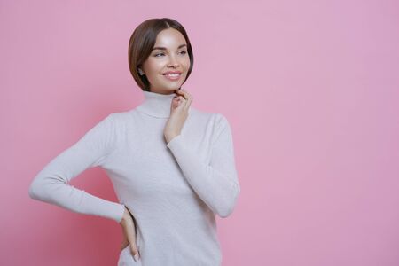 Beautiful tender young woman with toothy smile, looks aside pensively, dressed in casual white turtleneck, being in good mood, stands against pink background, blank free space for your promotionの写真素材